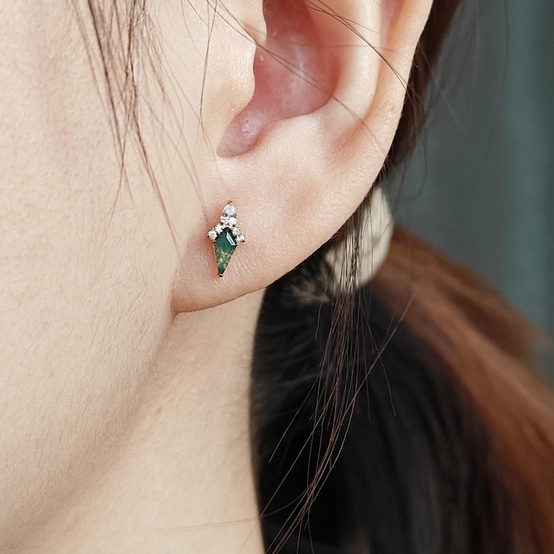 May include: A gold-colored stud earring with a green, diamond-shaped stone and small white accent stones. The earring is worn on an ear, showcasing its design and size. The earring's design is geometric and elegant.