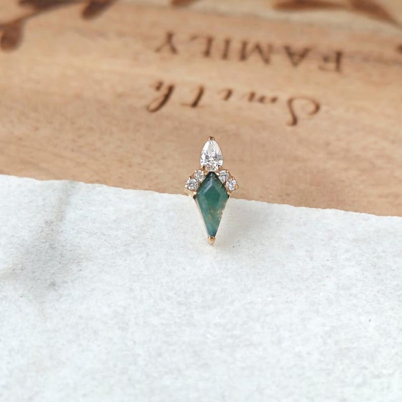 May include: A gold-toned earring with a geometric design. The earring features a teardrop-shaped clear stone at the top, a cluster of small clear stones, and a green, diamond-shaped stone at the base. The earring is set against a light-colored background.