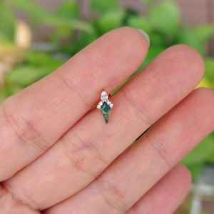 May include: A small, gold-colored earring with a green, diamond-shaped stone at the center. The earring is adorned with small, clear gemstones at the top. The design is delicate and elegant, suitable for ear piercings.