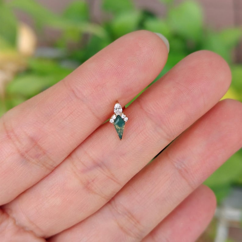 May include: A small, gold-colored earring with a green, diamond-shaped stone at the center. The earring is adorned with small, clear gemstones at the top. The design is delicate and elegant, suitable for ear piercings.