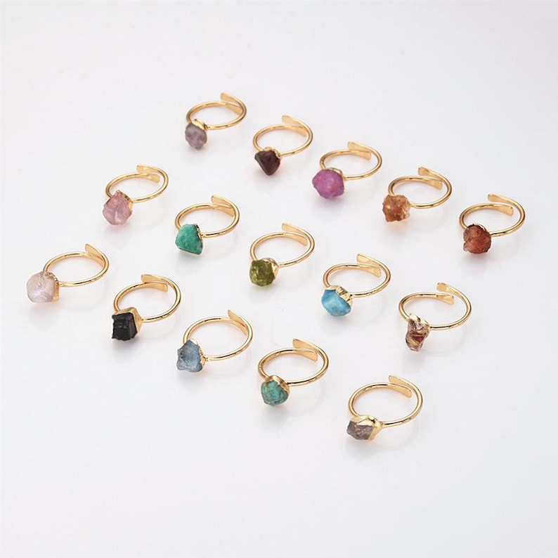 May include: A collection of adjustable gold-tone rings, each featuring a raw, uncut gemstone. The gemstones vary in color, including pink, green, blue, and brown. The rings are displayed on a white surface.