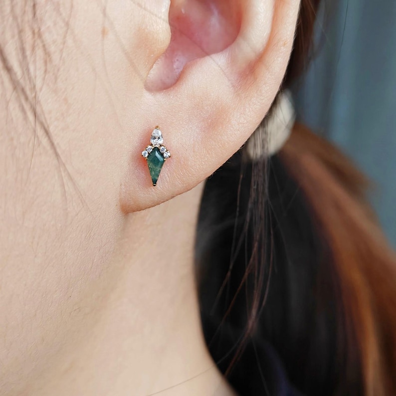 May include: A gold-toned earring with a green gemstone in a kite shape, accented with small, clear crystals. The earring is worn on an ear, showcasing its design and sparkle. The jewelry piece is a stud earring.