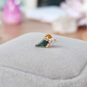 May include: A gold stud earring featuring a geometric design with a dark green, emerald-cut stone and several small, clear stones. The earring is set on a gray velvet surface.