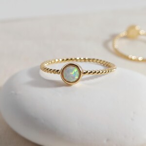 Puede incluir: Un anillo de oro con una banda de estilo cuerda trenzada y una piedra preciosa de &oacute;palo redonda engastada. El &oacute;palo muestra destellos iridiscentes de luz verde y azul. Un segundo anillo de oro desenfocado es visible en la esquina superior derecha.