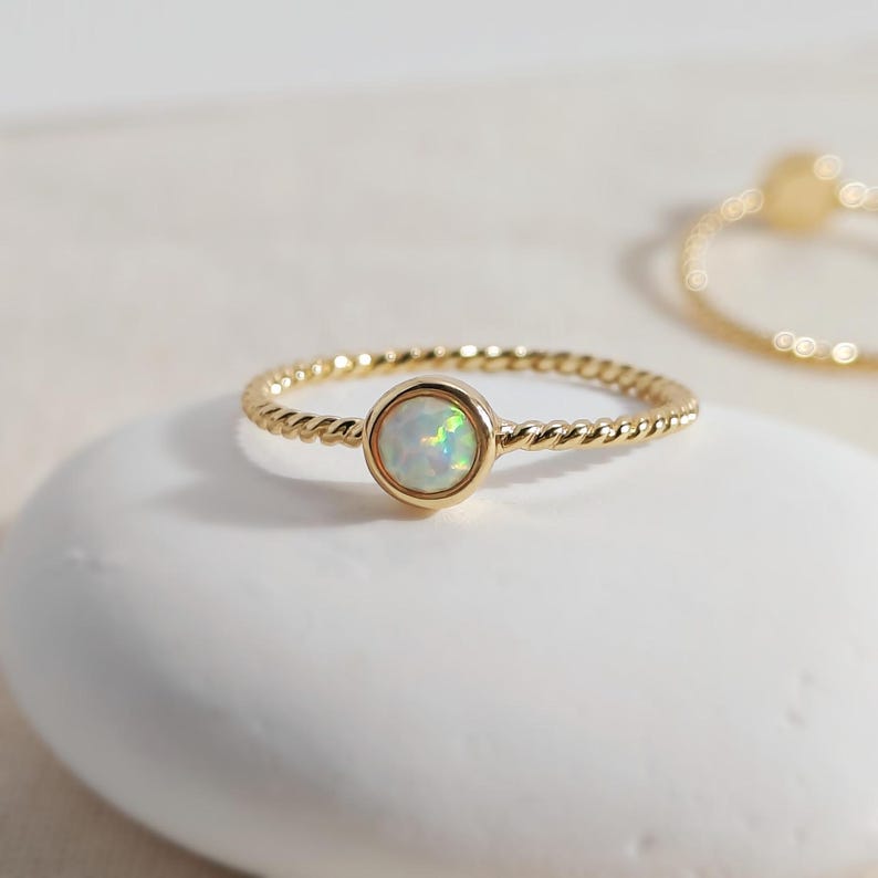 Puede incluir: Un anillo de oro con una banda de estilo cuerda trenzada y una piedra preciosa de &oacute;palo redonda engastada. El &oacute;palo muestra destellos iridiscentes de luz verde y azul. Un segundo anillo de oro desenfocado es visible en la esquina superior derecha.