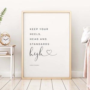 May include: A white framed print with a black and white quote that reads "Keep your heels, head and standards high" with a small heart symbol and the text "Coco Chanel" at the bottom.