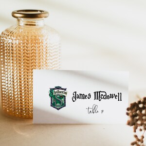 May include: A clear glass bottle with a gold-colored lid and a textured surface. A white card features the Slytherin crest and the name "James McDowell" with "table 7" below.