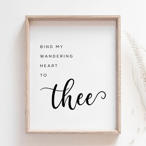 May include: A framed print with the words "Bind My Wandering Heart To Thee" in black script. The print is in a light-colored wooden frame, set against a white background. A sprig of dried grass is visible on the right side.