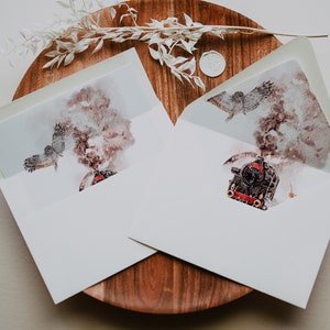 May include: Two white envelopes with watercolor illustrations of a steam train and an owl in flight. The envelopes are open, revealing the artwork inside. The envelopes are on a wooden surface with decorative elements.