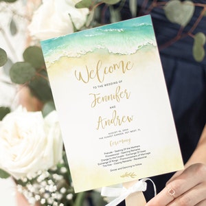 Puede incluir: Un programa de boda con una escena de playa en acuarela. La escritura dorada dice "Bienvenida a la boda de Jennifer y Andrew". Los detalles de la ceremonia se enumeran a continuación. El programa se sostiene con un palo de madera y una cinta blanca.