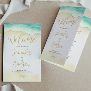 May include: Welcome sign for a wedding with a watercolor background of ocean waves and a sandy beach. The text reads "Welcome to the wedding of Jennifer and Andrew" with the date and location below.