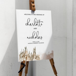 May include: A white wedding welcome sign on a wooden easel. The sign reads "Welcome to the Wedding of Charlotte and Nicholas" with the date and location below. A watercolor image of a castle is at the bottom.