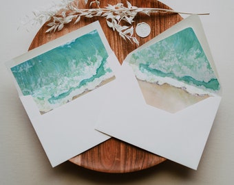 May include: Two white envelopes with a watercolor design of ocean waves and a sandy beach on the front. The envelopes are on a wooden plate.