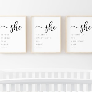 May include: Three framed prints with the word "she" in elegant script. Each print features a different inspirational quote in a minimalist black and white design. The frames are a light wood color.