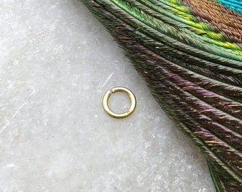 May include: A small, gold-colored hoop earring. The earring is lying on a white surface with a peacock feather in the background.