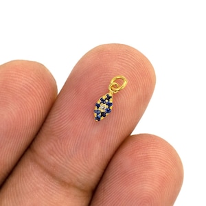 Könnte beinhalten: Goldener Anhänger mit einem blauen und weißen bösen Auge. Der Anhänger ist tropfenförmig und hat oben eine kleine Öse zum Anbringen an einer Halskette oder einem Armband.