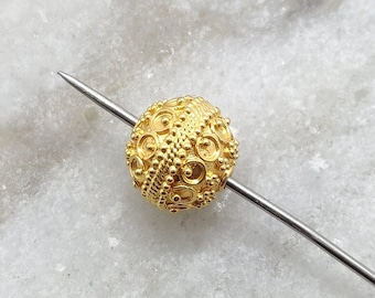 May include: A gold-colored metal bead with a detailed, intricate design. The bead is round and has a textured surface.