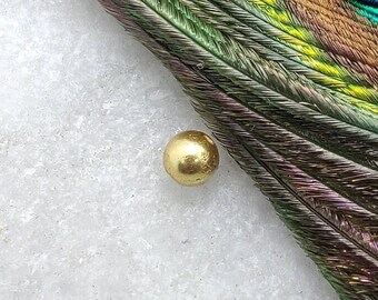 May include: A single gold bead resting on a white surface with a peacock feather in the background.