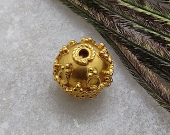 May include: A gold-colored bead with a detailed design and a hole in the center. The bead is on a white surface with a green and brown feather in the background.