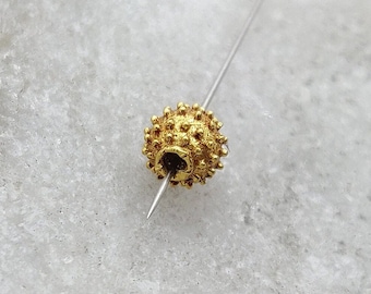May include: A gold-colored bead with a textured surface, resembling a tiny sea urchin, is threaded on a thin silver wire.