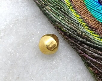 May include: A small, round, gold-colored bead on a white marble surface. The bead is slightly irregular in shape and has a matte finish. A peacock feather with iridescent green, blue, and brown colors is partially visible in the background.