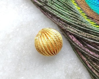 May include: A gold-colored, round bead with a textured surface. The bead is sitting on a white marble surface.