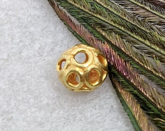 May include: A gold-colored metal bead with a geometric design. The bead has a circular shape with multiple open circles around the outside. The bead is sitting on a white surface.