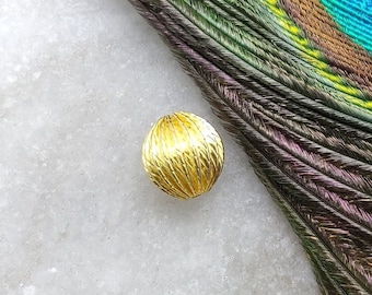 May include: A small, round, gold-colored bead with a textured surface. The bead is sitting on a white marble surface.