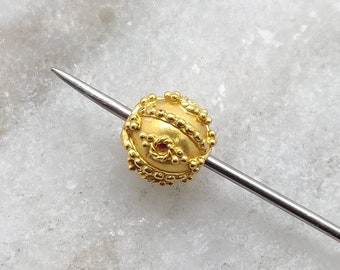 May include: A gold-colored bead with a textured surface and a small hole in the center. The bead is on a thin metal rod.