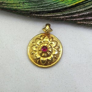 May include: A gold-colored pendant with a floral design and a small, round, red gemstone in the center. The pendant has a loop at the top for a chain. The background includes a peacock feather and a white surface.