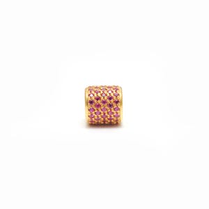 May include: A gold-colored cylindrical bead encrusted with numerous small, round, pink gemstones. The bead has a polished finish and is set against a white background. The gemstones are closely packed together, creating a textured surface.