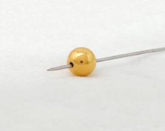 May include: A gold-colored, spherical bead pierced by a thin, silver-colored metal pin. The bead has a smooth, reflective surface. The pin is centered through the bead, creating a simple, elegant design. The background is a soft, white fabric.