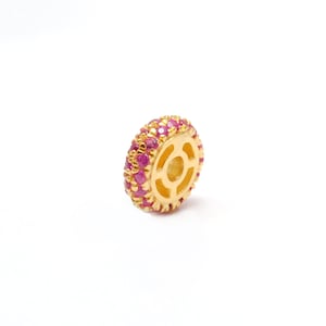 May include: A gold-colored bead with a wheel-like design. The bead is encrusted with small, round, pink gemstones around the outer edge. The center of the bead has a circular opening with a geometric pattern.