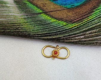May include: A gold-colored clasp with a red gemstone in the center. The clasp is shaped like an infinity symbol.