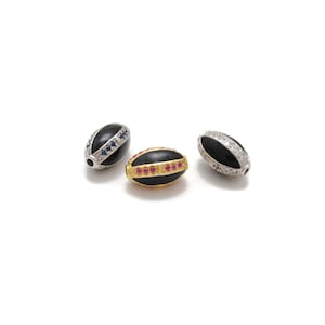 Puede incluir: Tres cuentas ovaladas con esmalte negro y detalles de piedras preciosas. Una cuenta tiene una banda dorada con piedras rojas, las otras dos tienen bandas plateadas con piedras azules y transparentes. Sobre fondo blanco.