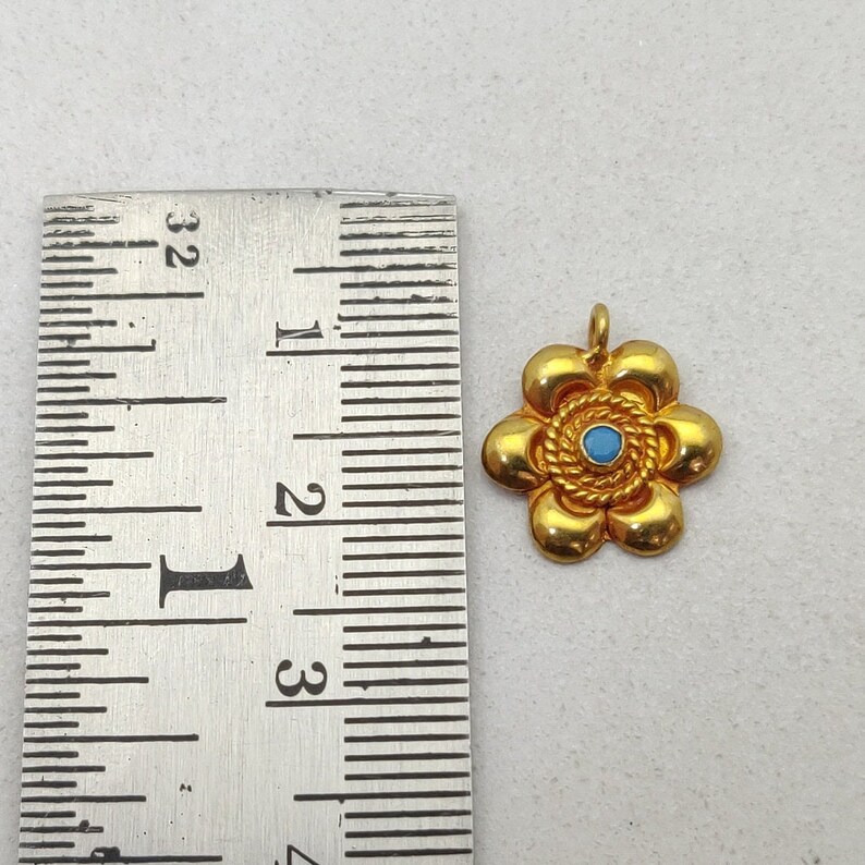 May include: A gold-colored flower charm with a blue center stone. The charm is about 1 inch in diameter and is shown next to a ruler.