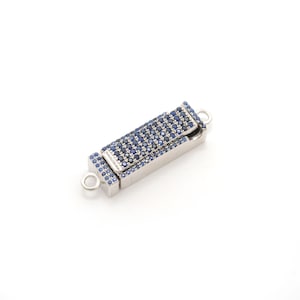 Peut inclure: Un fermoir rectangulaire argenté avec un dessus articulé, incrusté de petites pierres précieuses bleu foncé. Le fermoir comporte deux petites boucles à chaque extrémité pour la fixation. Les pierres précieuses sont disposées en motif dense.