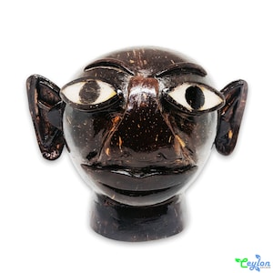 May include: A hand-carved wooden sculpture of a face with large ears, a prominent nose, and exaggerated eyes. The sculpture is made from dark brown coconut shell and has a distressed finish.