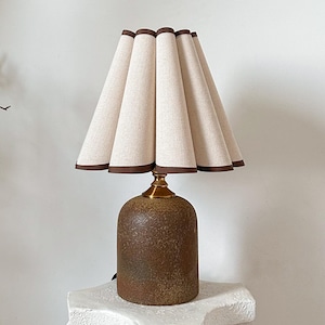 Könnte beinhalten: Dekorative Tischlampe mit beigefarbenem, plissiertem Schirm, braun eingefasst. Die Lampe hat einen strukturierten braunen Sockel und einen Messinghals. Ein stilvolles Wohnaccessoire.