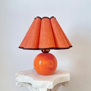 May include: A table lamp featuring a glossy, spherical orange base and a pleated, textured orange fabric lampshade. The shade is finished with a dark brown trim along the bottom edge, and the lamp is connected by a small, metallic gold-toned neck.
