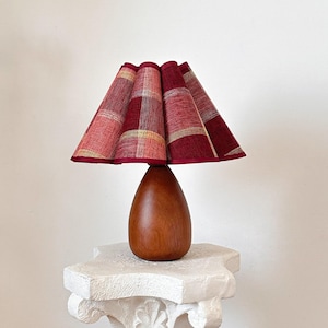 Op de afbeelding: Een kleine tafellamp met een rood en goud geruite lampenkap. De voet is glad, druppelvormig hout. De lamp staat op een witte, decoratieve sokkel. Een zwart snoer loopt van de basis.