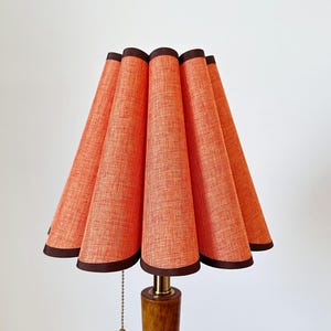 Puede incluir: Una lámpara de mesa con una pantalla plisada de color naranja texturizado y bordes en marrón oscuro. La lámpara cuenta con una base cilíndrica de madera y un interruptor de cadena metálica que cuelga bajo la pantalla.