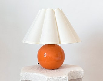 Puede incluir: Una lámpara de mesa con una base esférica de cerámica naranja brillante y una pantalla de tela plisada en color blanco roto. La pantalla tiene una forma cónica ancha con pliegues verticales, sobre una base de esfera naranja lisa.