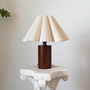 Könnte beinhalten: Ein weißer, gefalteter Lampenschirm sitzt auf einem Lampenfuß aus Holz. Die Lampe steht auf einem weißen Sockel mit verzierten Details.