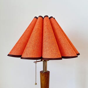 Puede incluir: Una lámpara de mesa con pantalla de tela naranja plisada y ribete marrón oscuro. La lámpara tiene una cadena de latón y una base cilíndrica de madera. La pantalla presenta un diseño de borde festoneado y una textura tejida visible.