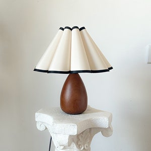 May include: A white lampshade with a black trim sits atop a wooden base. The lamp is on a white pedestal.