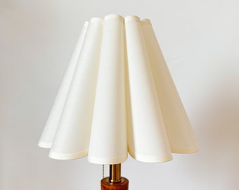 Puede incluir: Una lámpara de mesa con una pantalla de tela plisada en color blanco roto con borde festoneado. La base de la lámpara está hecha de una pieza cilíndrica de madera pulida con un cuello de metal color latón y un interruptor de cadena.