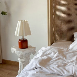 May include: A white lampshade with a ruffled edge sits on a white pedestal table. The lamp has a wooden base and is next to a white bed with a woven headboard.