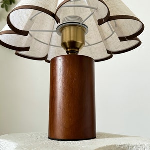 K&ouml;nnte beinhalten: Eine Tischlampe aus Holz mit einem braunen Sockel und einem wei&szlig;en Stoffschirm mit gewellten R&auml;ndern.