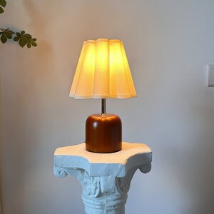 May include: A brown wooden table lamp with a white pleated lampshade. The lamp is turned on and the light is shining through the shade.